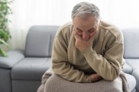 A distressed older man sits at the edge of his sofa lost in thought